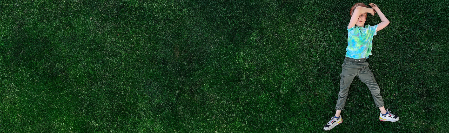 Ein Kind liegt in der Wiese, den linken Arm über dem Kopf, der rechte Arm liegt neben dem Kopf in der Wiese. Das Kind Blickt in die Kamera.